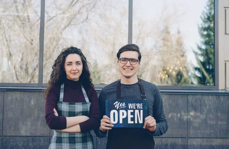 Two small business owners in aprons holding a “Yes, we’re open” sign outside their shop