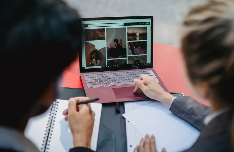 Two people reviewing a grid of photos on a laptop while taking notes, like approving a monthly social media proof